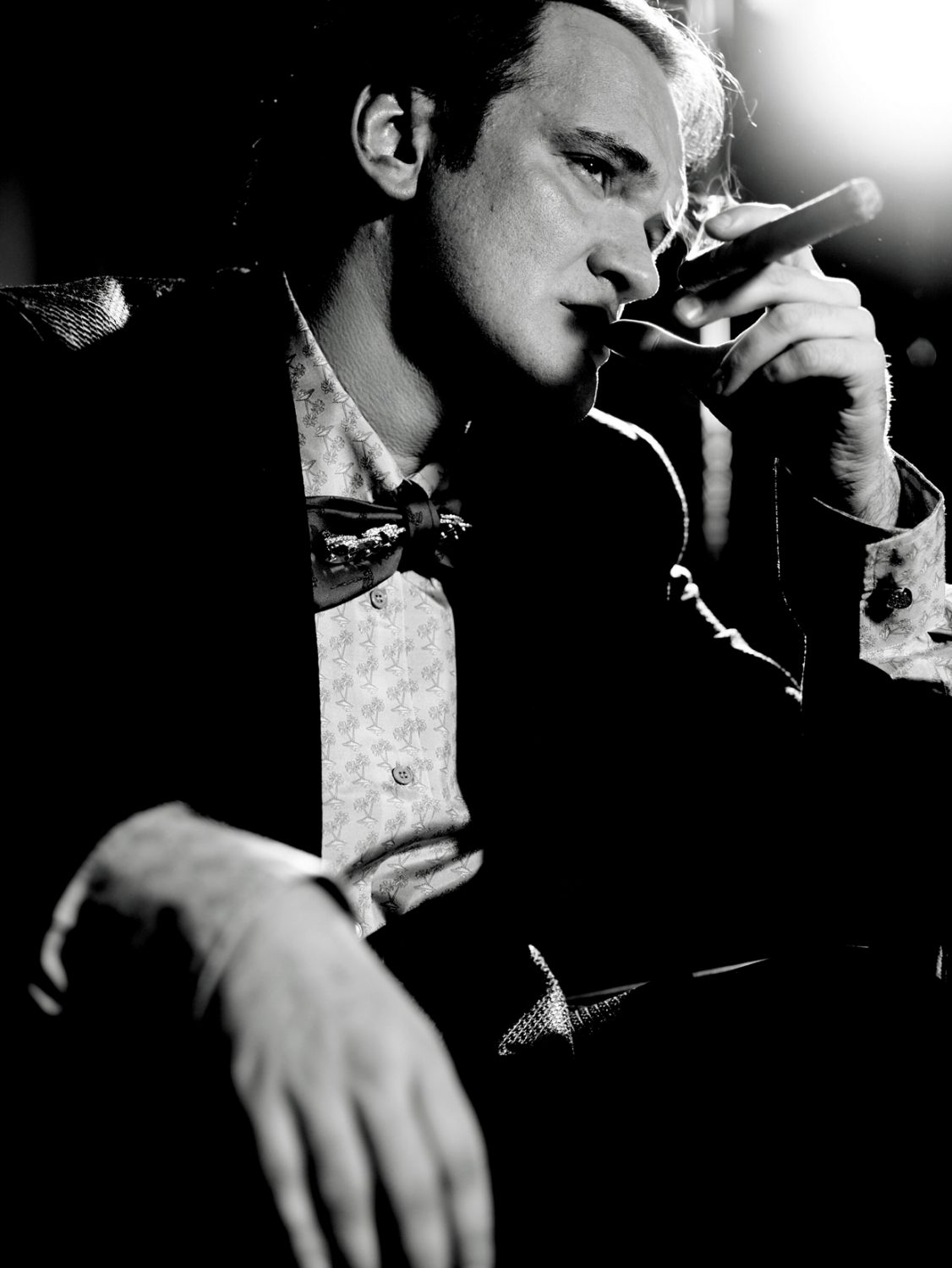 Black and white portrait of a man with dark hair, wearing a patterned shirt and bow tie, smoking a cigar in dark, moody lighting.