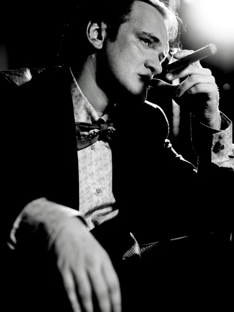 Black and white portrait of a man with dark hair, wearing a patterned shirt and bow tie, smoking a cigar in dark, moody lighting.