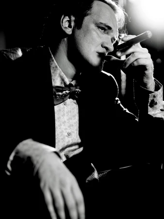 Black and white portrait of a man with dark hair, wearing a patterned shirt and bow tie, smoking a cigar in dark, moody lighting.