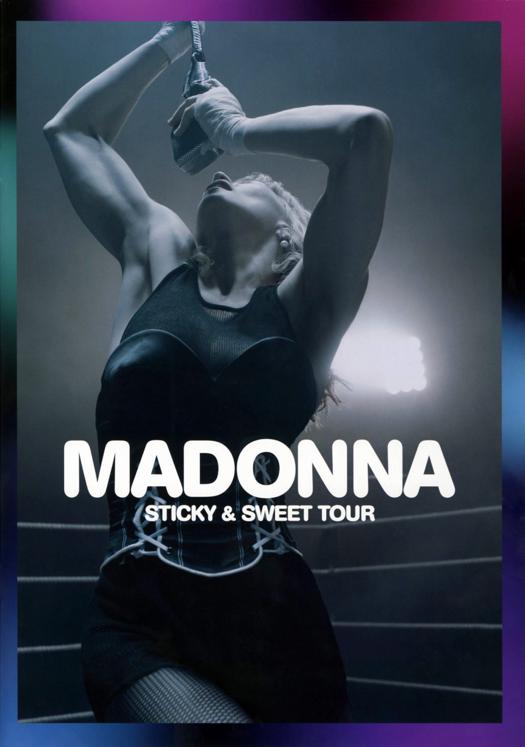 Madonna in a black corset and fishnets holds a microphone overhead in a moody, theatrical concert lighting setting against a faded background.