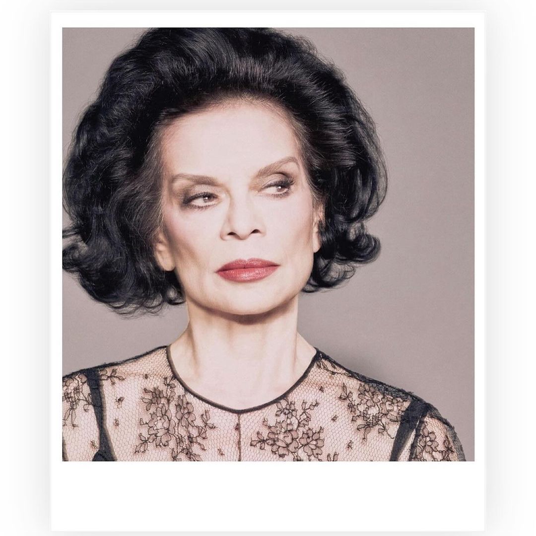 Elegant portrait of an adult woman with dark curly hair in black floral lace top against a gray backdrop, photographed indoors.