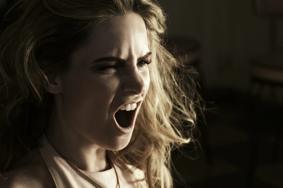 Close-up portrait of a young adult female with wavy blonde hair, shouting with an open mouth and an intense facial expression in dim light.