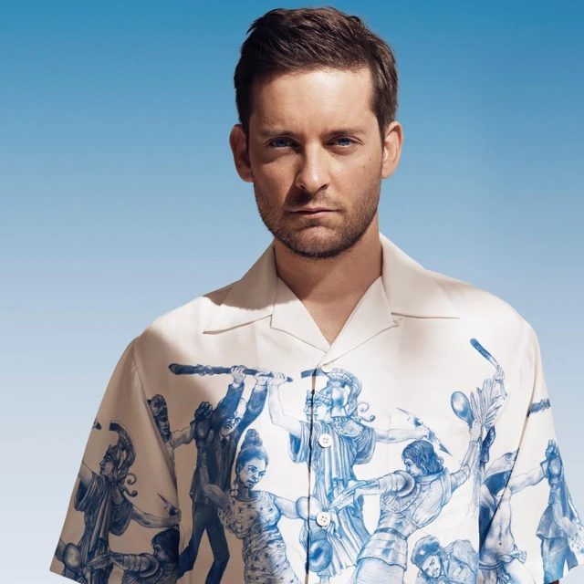 Close-up studio portrait of a man wearing a printed white Prada shirt against a pale blue backdrop with soft, diffused lighting.