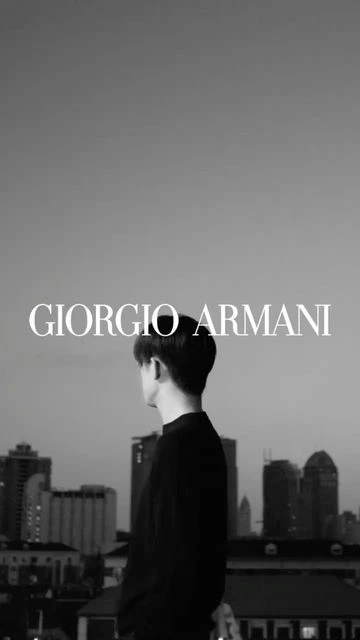 Black-and-white shot of a person looking at city buildings under a twilight sky, with 'GIORGIO ARMANI' text overlay.