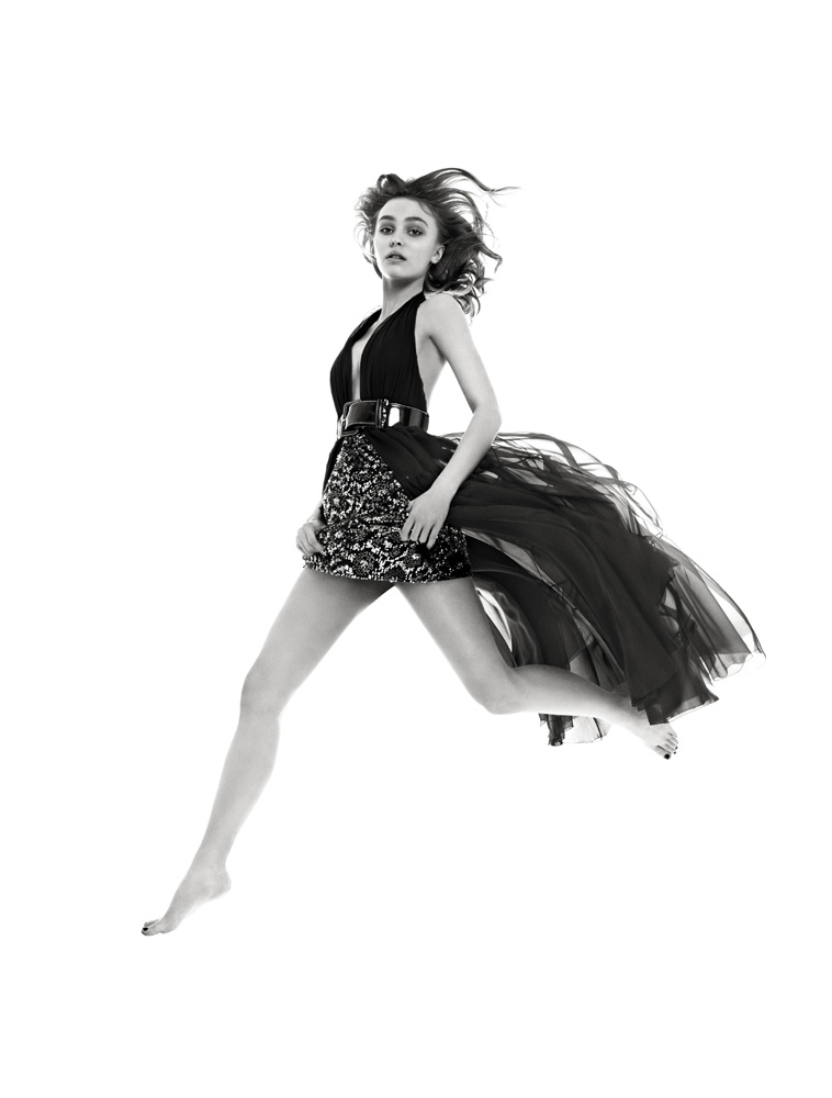 Black and white fashion portrait of a young adult woman leaping mid-air wearing a dress with a layered skirt against a white backdrop.