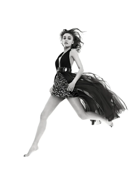 Black and white fashion portrait of a young adult woman leaping mid-air wearing a dress with a layered skirt against a white backdrop.