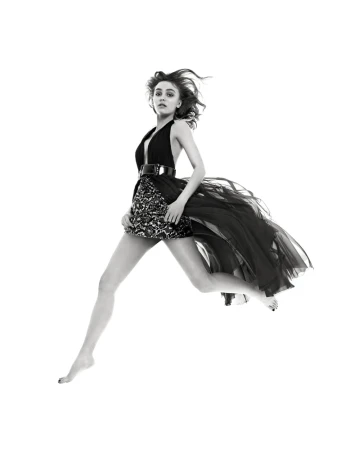 Black and white fashion portrait of a young adult woman leaping mid-air wearing a dress with a layered skirt against a white backdrop.