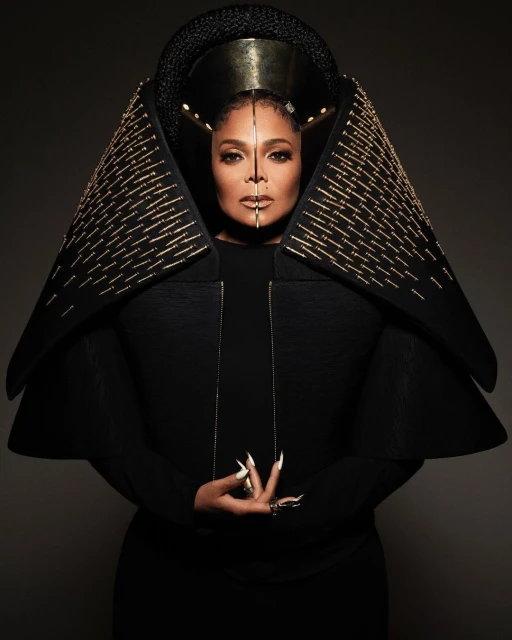 Portrait of Janet Jackson wearing an elaborate black and gold costume with long nails and rings, set against a dark grey background.