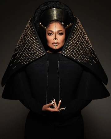 Portrait of Janet Jackson wearing an elaborate black and gold costume with long nails and rings, set against a dark grey background.