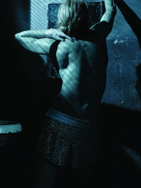 Back view of a woman with blonde hair, wearing sparkly shorts, in a dimly lit room with a window casting shadows across her back.