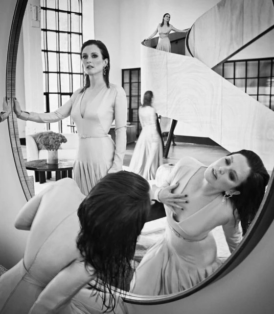 Black and white portrait of a woman in a dress with multiple reflections, standing near a staircase. Indoor, elegant composition.