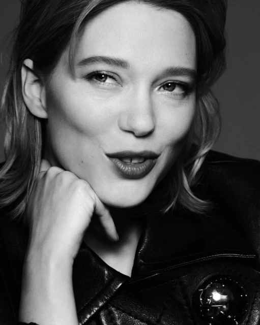 Black and white portrait of Léa Seydoux in a leather jacket, resting her chin on her hand, with a neutral background.