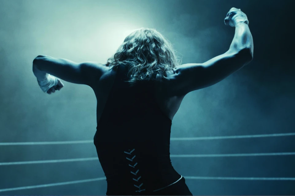 An athletic woman, seen from the back, stands in a boxing ring with arms raised, celebrating, dressed in athletic wear with wrapped hands.