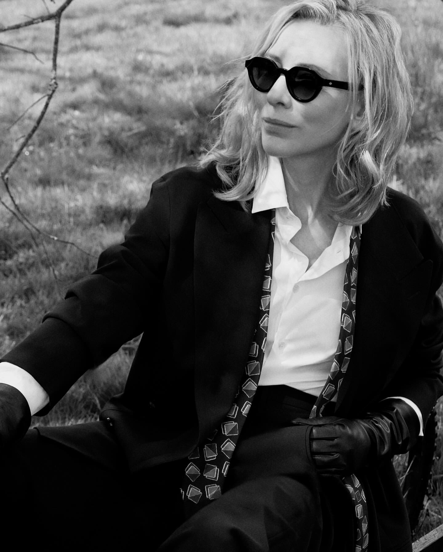 Black-and-white portrait of a blonde adult female in a suit and sunglasses, seated in a grassy outdoor setting. Editorial photography.