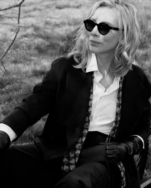 Black-and-white portrait of a blonde adult female in a suit and sunglasses, seated in a grassy outdoor setting. Editorial photography.