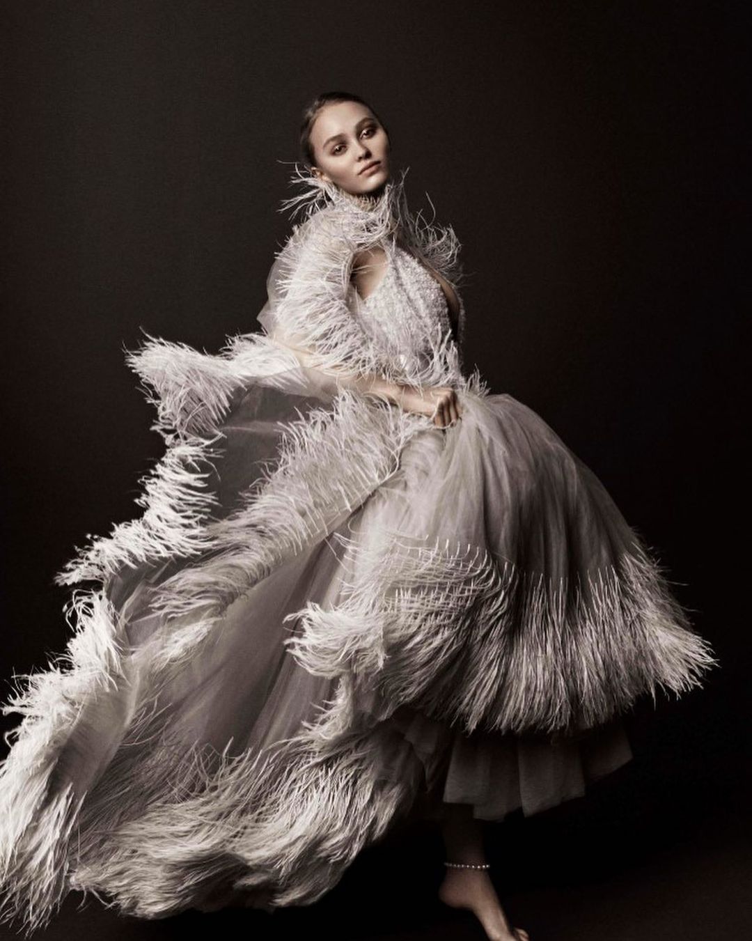 Elegant woman in an elaborate white feathered gown strikes a pose in a studio, creating a visually striking fashion portrait.