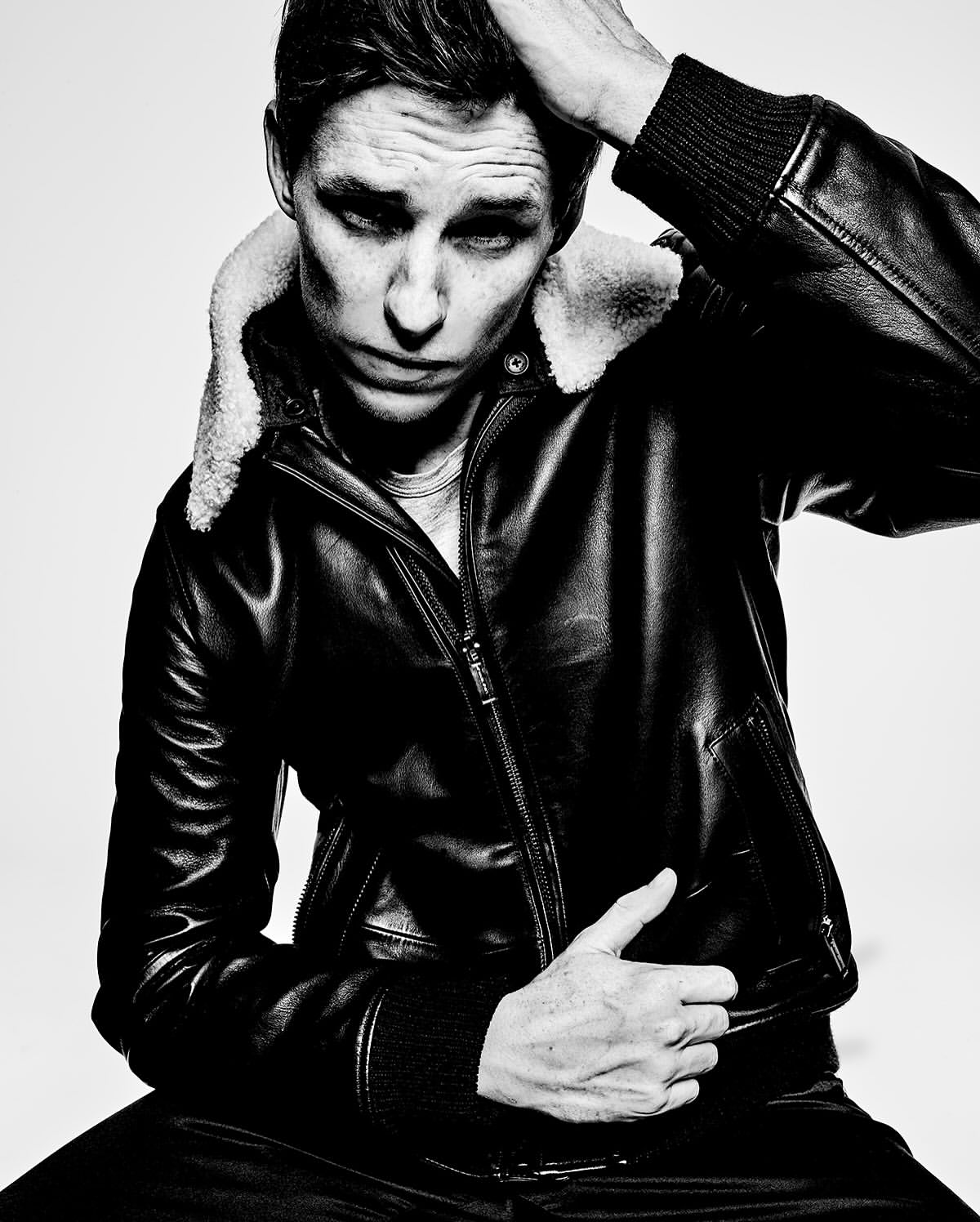 Portrait of a dark-haired man wearing a black leather jacket with a fur collar, hand on his head, against a light background.