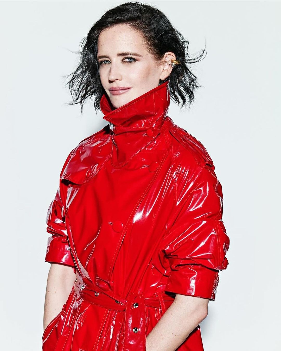 Close-up studio shot of Eva Green wearing a red, patent leather coat. Her dark hair is styled short, and she wears an ear cuff.