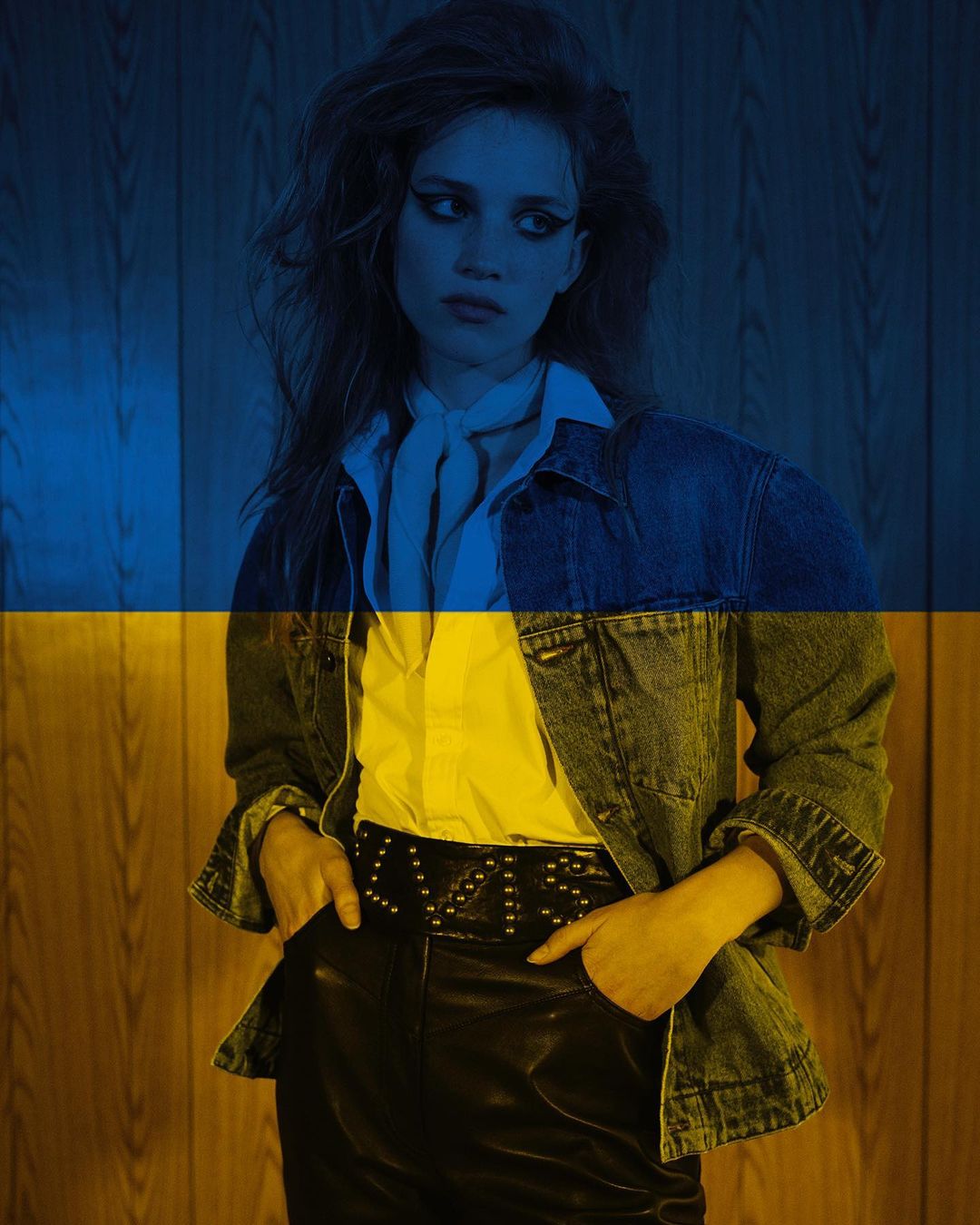 Portrait of a young adult female model in a denim jacket, white shirt, and black pants, with a yellow and blue flag overlay.