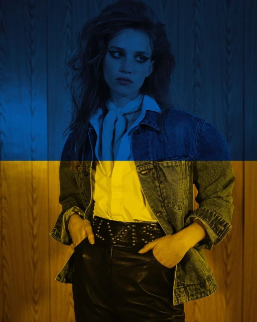 Portrait of a young adult female model in a denim jacket, white shirt, and black pants, with a yellow and blue flag overlay.