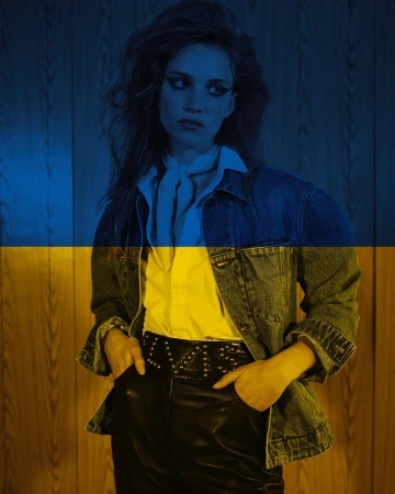 Portrait of a young adult female model in a denim jacket, white shirt, and black pants, with a yellow and blue flag overlay.