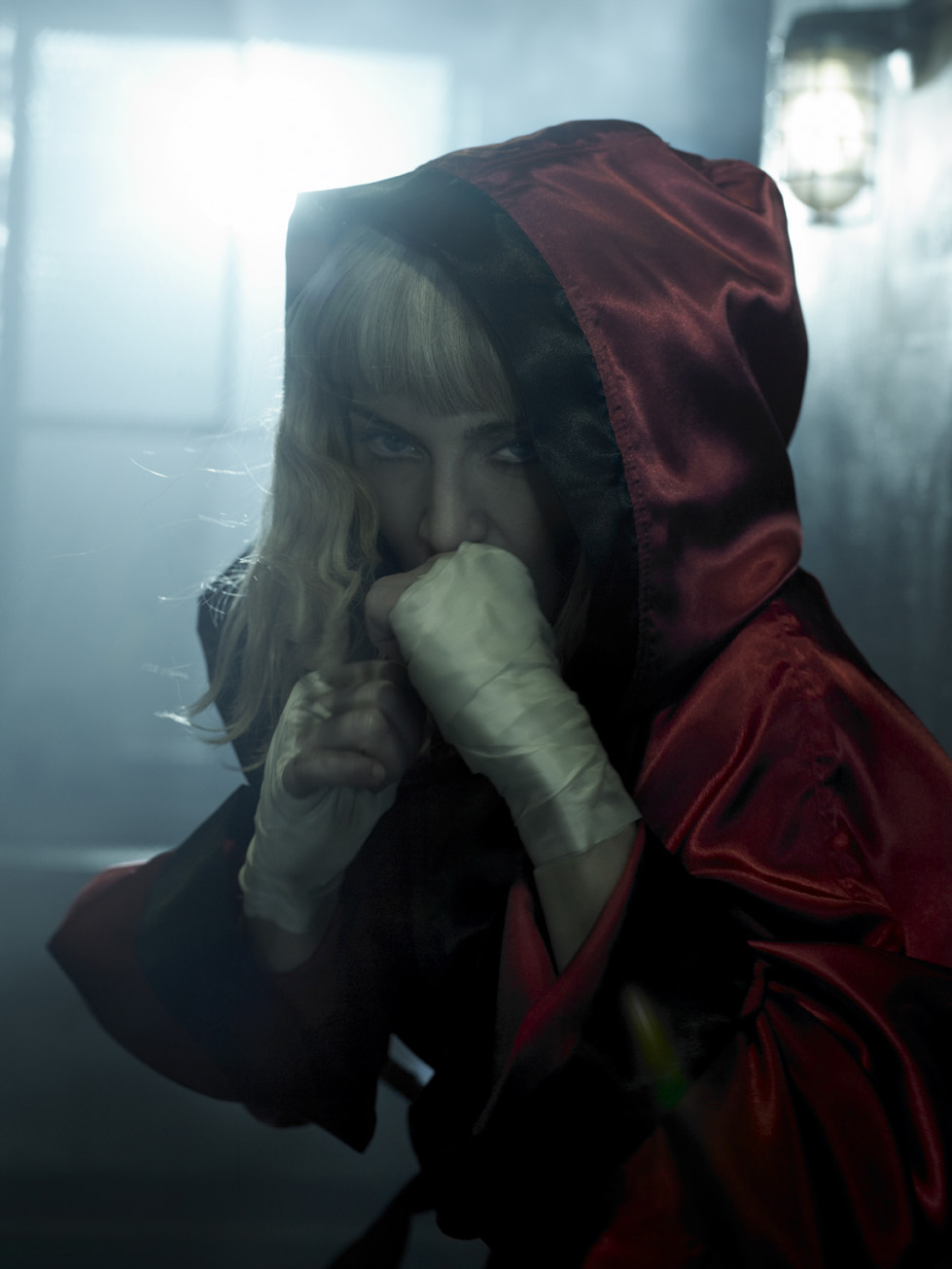 Woman with blonde hair in a red hooded robe and hand wraps posing in a dimly lit indoor setting with a moody atmosphere.