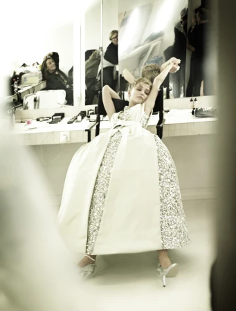 A young woman in a voluminous white and silver sequin ballgown stretching her arms in a dressing room mirror reflection.