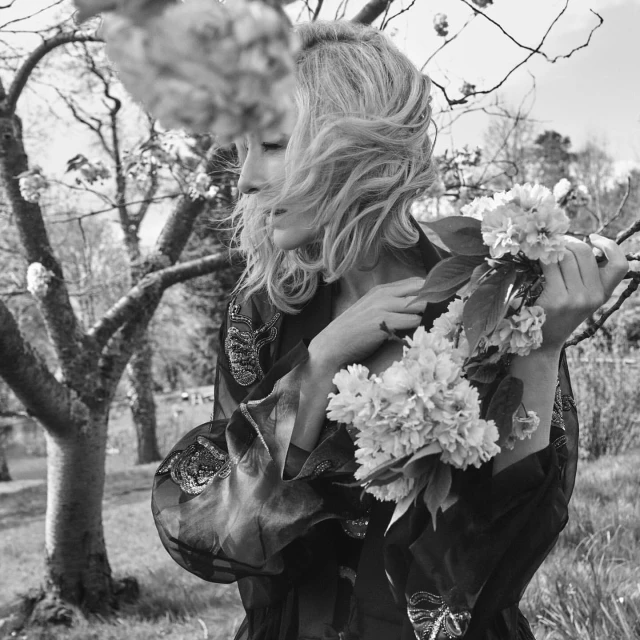 Black and white portrait of a blonde woman with a sheer patterned robe holding a flowering branch in an outdoor, grassy setting.