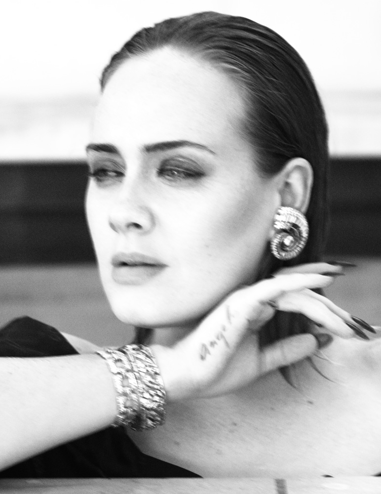 Black and white portrait of a stylish woman with slicked-back hair, wearing large earrings and a bracelet, with a tattoo on her hand.