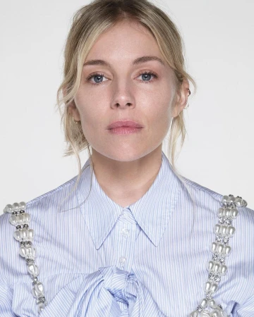 Portrait of Sienna Miller wearing a light blue pinstripe shirt with pearl-embellished straps against a plain white background.