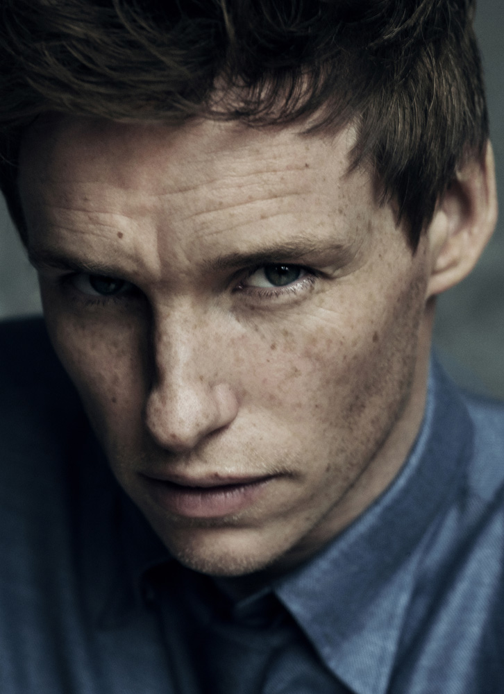 Close-up portrait of an adult male with freckles wearing a blue button-down shirt against a neutral gray backdrop in studio lighting.