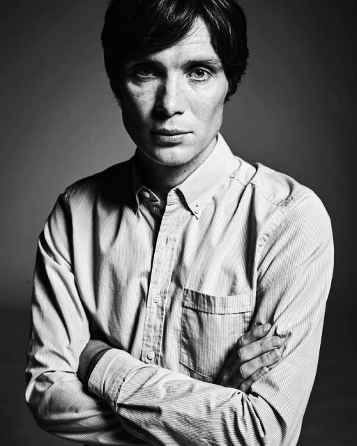 Black-and-white portrait of Cillian Murphy in a striped dress shirt with arms crossed, against a neutral grey backdrop, captured in a studio setting.