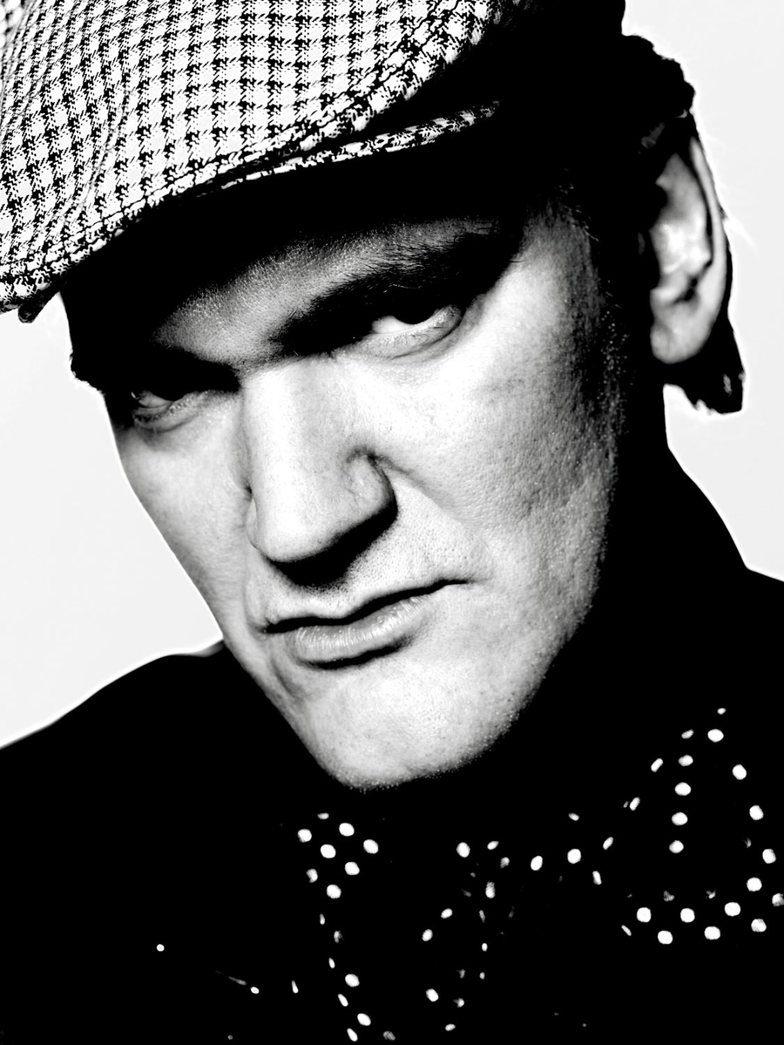 Monochrome portrait of an adult male wearing a newsboy cap and polka dot bow tie against a light background in a studio setting.