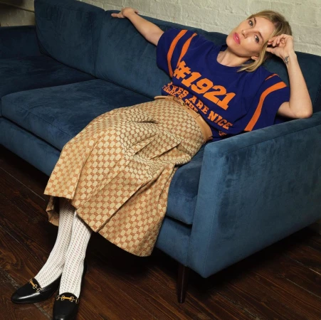 Woman with blonde hair seated on a dark blue velvet sofa wearing a blue t-shirt and patterned beige skirt