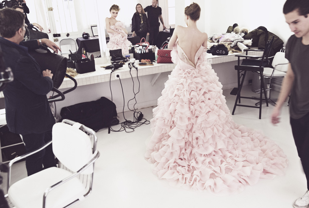 Model wearing a voluminous pale pink ruffled gown with a train in a backstage dressing room filled with crew members.