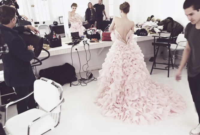 Model wearing a voluminous pale pink ruffled gown with a train in a backstage dressing room filled with crew members.