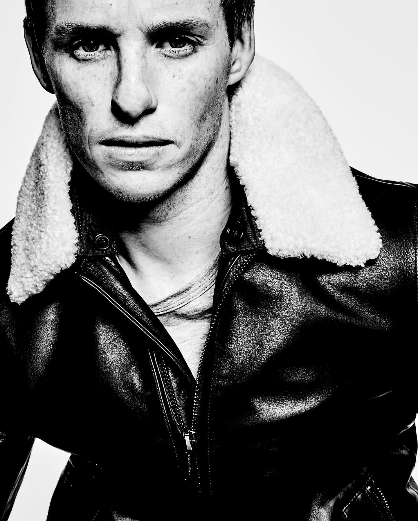 Headshot of Eddie Redmayne in black and white, wearing a leather jacket with a cream shearling collar, gazing intensely at the camera.