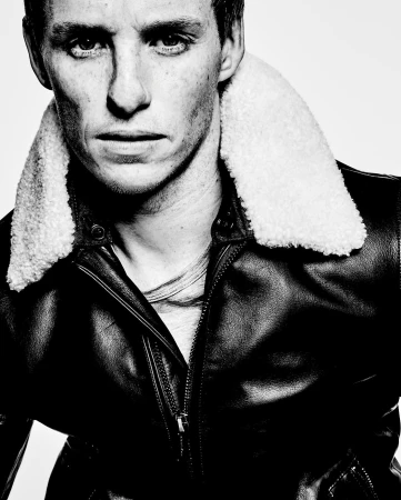 Headshot of Eddie Redmayne in black and white, wearing a leather jacket with a cream shearling collar, gazing intensely at the camera.