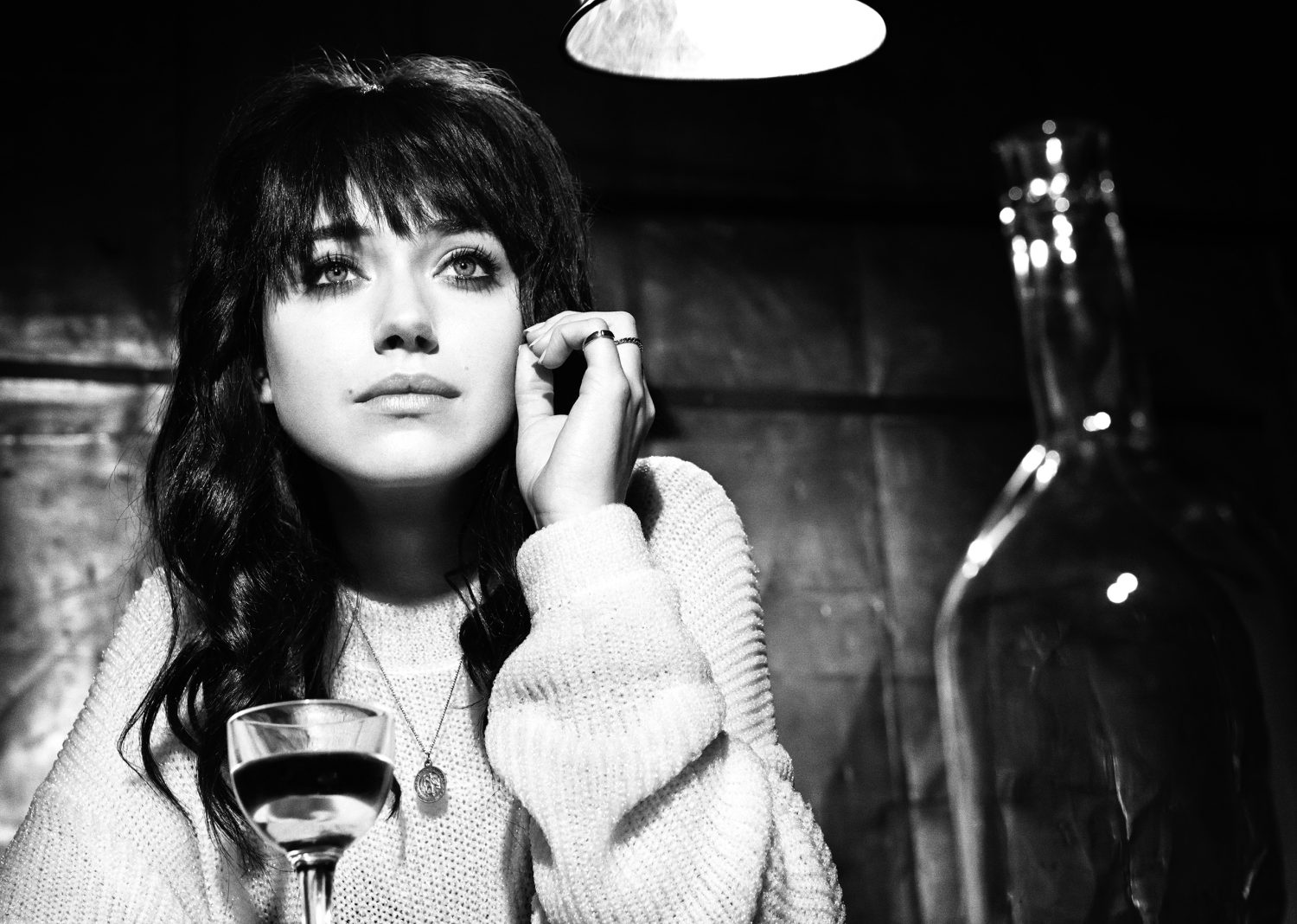 Black and white portrait of a young woman with dark hair, wearing a sweater, sitting at a table with a wine glass.