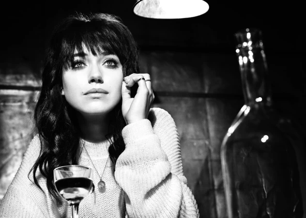 Black and white portrait of a young woman with dark hair, wearing a sweater, sitting at a table with a wine glass.
