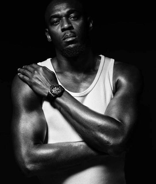 Black and white studio portrait of a muscular adult male wearing a white tank top with arms crossed, showcasing a watch, set against a dark background.