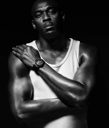 Black and white studio portrait of a muscular adult male wearing a white tank top with arms crossed, showcasing a watch, set against a dark background.