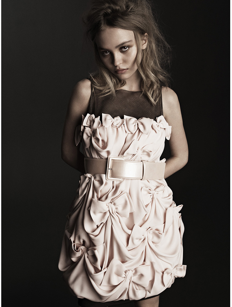 Lily-Rose Depp wearing a pale pink bow-adorned dress with a wide belt against a dark background