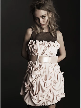 Lily-Rose Depp wearing a pale pink bow-adorned dress with a wide belt against a dark background
