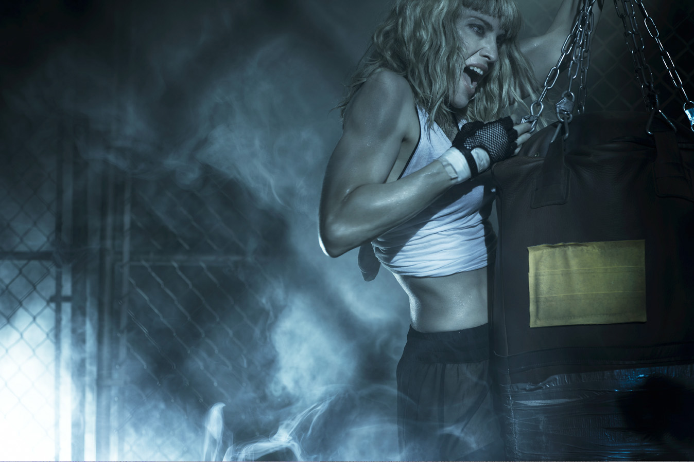 Muscular blonde woman in workout gear punches a punching bag in a boxing ring, illuminated by dramatic lighting and surrounded by fog.