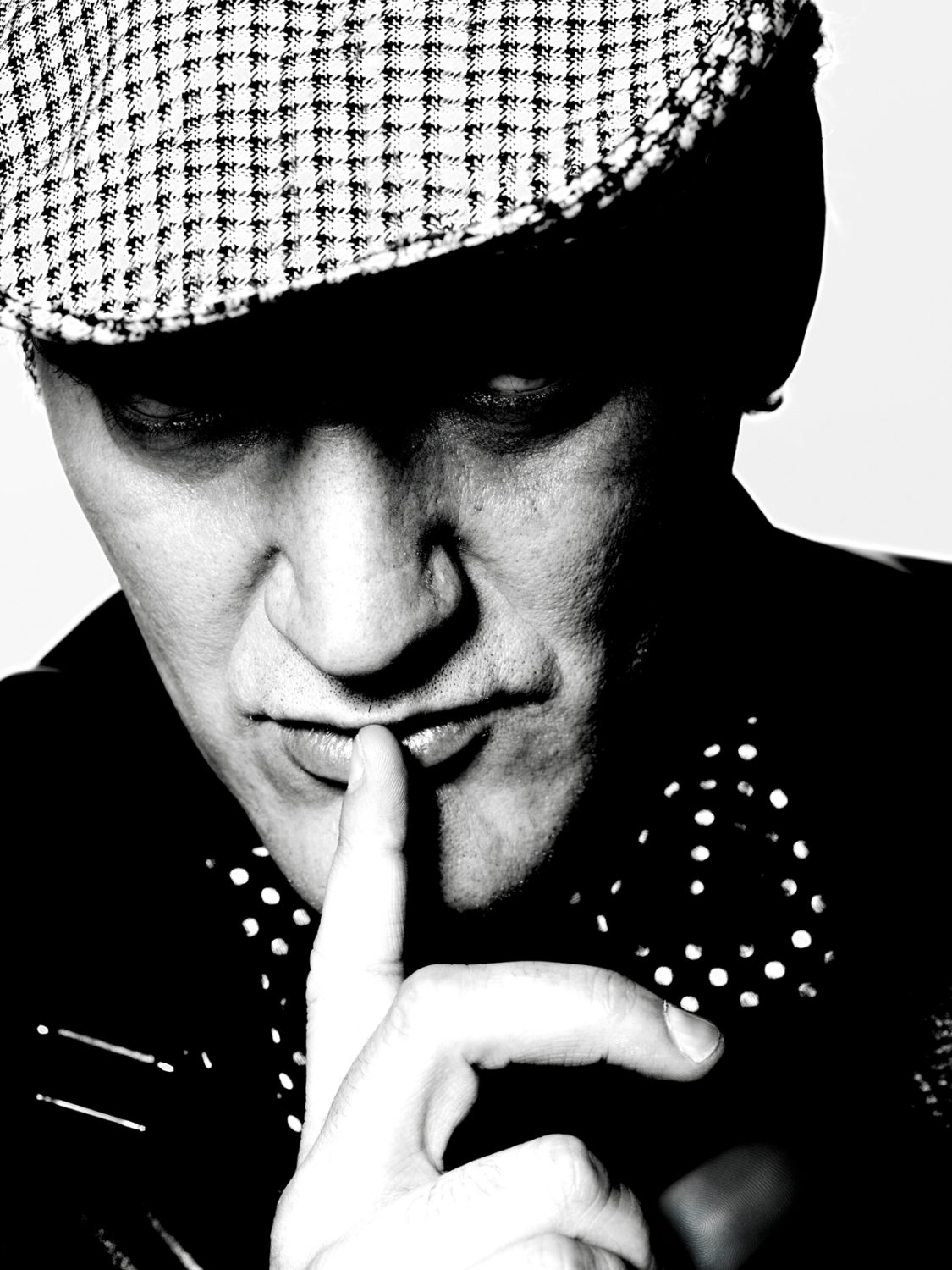 Black-and-white portrait of an adult male with finger to lips, wearing a newsboy cap and polka dot shirt against a light background.