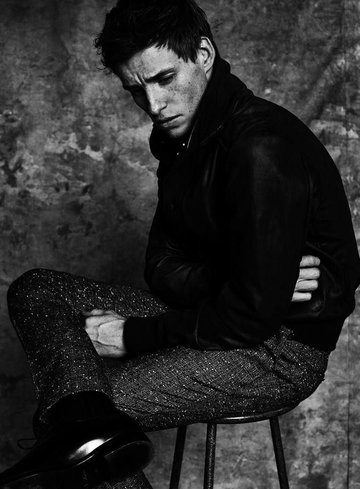 Black and white portrait of a man in a leather jacket, sitting on a stool in a studio setting, showcasing a somber mood.