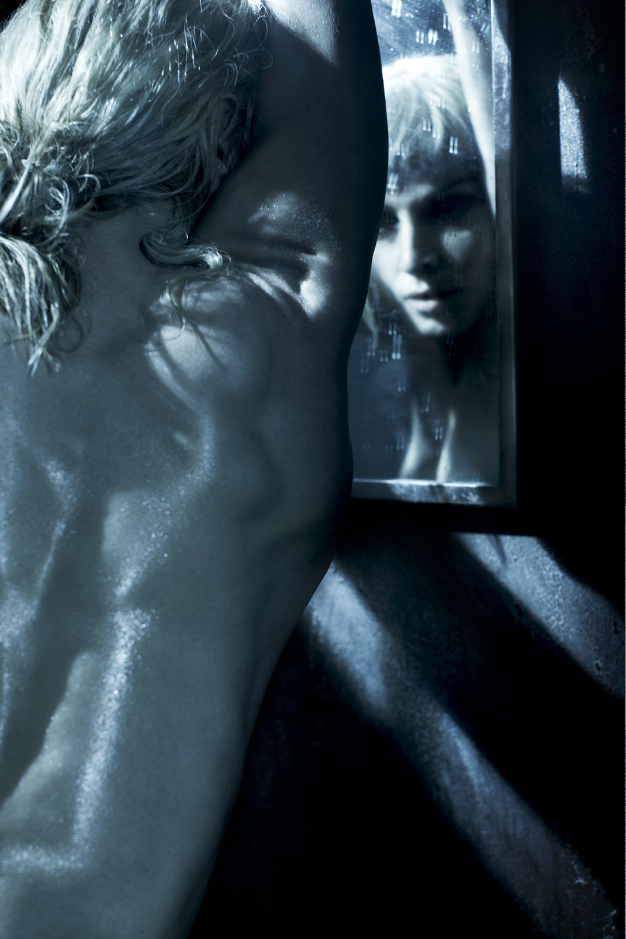 Woman looking in a mirror with cool-toned lighting and shadows; bare shoulders are in the foreground of the shot.