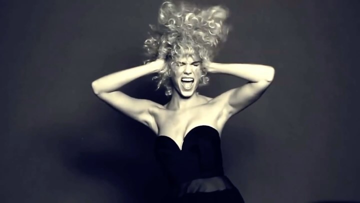 Black and white studio shot of a woman with blonde curly hair, wearing a black dress, with her hands near her head, mouth open.
