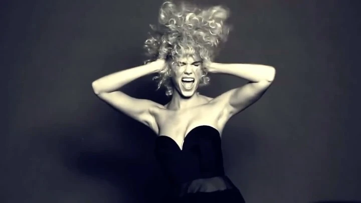 Woman in a strapless black dress with voluminous curly blonde hair holding her head and screaming against a dark background.
